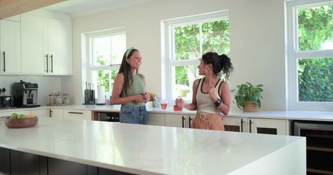 Diverse female friends chatting in bright modern kitchen holding pink drinks
