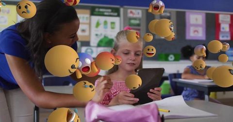 Teacher leaning over girl using tablet with floating emoji ar in classroom learning