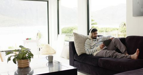 Indian man relaxing on modern sofa with tablet and headphones, bright lakeview living room