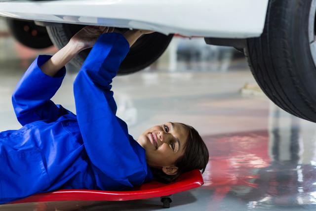 Female mechanic lying under a car, working on vehicle repair in a service center. She is wearing a blue jumpsuit, focused on the task. Ideal for promoting automotive services, gender diversity in technical professions, and mechanic training programs.