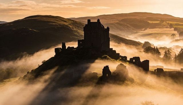Medieval castle silhouette on misty rolling hills at golden hour dawn, fog-wrapped ruins