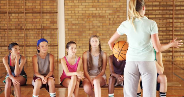 Diverse teenage basketball team listening attentively to coach during practice in an indoor gym. Ideal for usage in content related to sports coaching, team building, youth sports, physical education, and exercise programs.