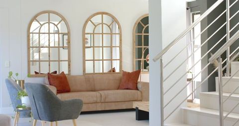 Sunlit minimalist living room featuring tan sofa, rust pillows and arched window mirrors