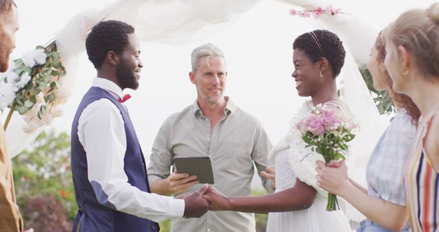 Diverse couple joyfully holding hands during an outdoor wedding ceremony surrounded by guests and an officiant. Perfect for use in advertising for wedding services, inclusive marriage events, or relationship blogs focusing on love and unity.