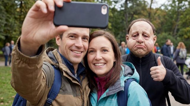 Smiling couple taking selfie in urban park while masked photobomber gives cheeky thumbs up