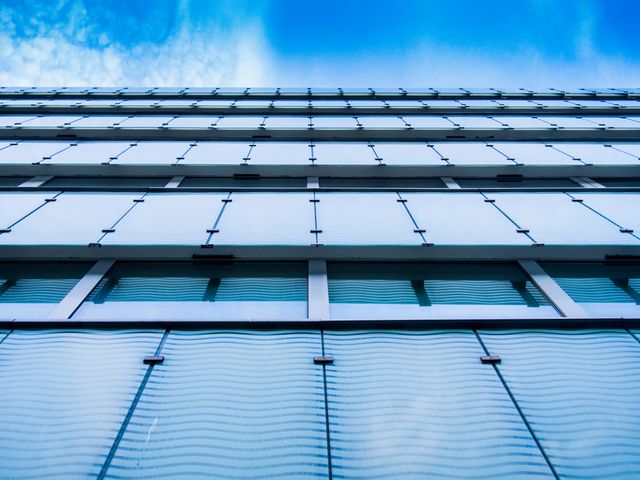 Image depicting the modern glass facade of a tall building viewed from below, creating a sense of height and perspective. Reflective windows contrast with the clear blue sky, emphasizing the architectural details. Ideal for use in magazines, blogs, urban development presentations, or as a background for corporate materials.