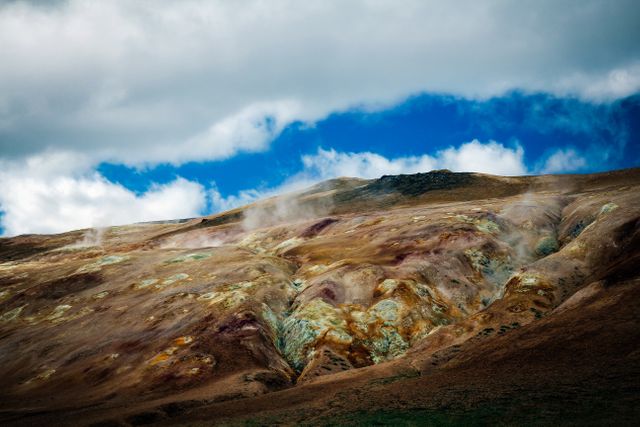 This image displays a vibrant geothermal landscape with steaming earth and volcanic hot springs under a dramatic, cloudy sky. With a backdrop of the partly cloudy blue sky, the colors of the earth and the steam create a striking and dynamic scene. Ideal for use in environmental articles, travel blogs, geology educational materials, and nature photography collections.