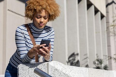 African american woman leaning on urban ledge checking smartphone in sunlit concrete plaza