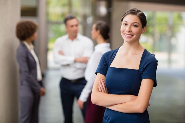 Confident businesswoman standing with arms crossed, smiling at camera, colleagues conversing in background. Ideal for use in business, leadership, teamwork, and corporate success themes. Suitable for websites, presentations, and promotional materials highlighting professional environments and career growth.