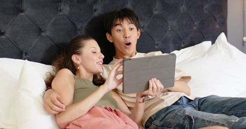 Couple lounging on bed sharing tablet, surprised reaction, cozy bedroom lifestyle
