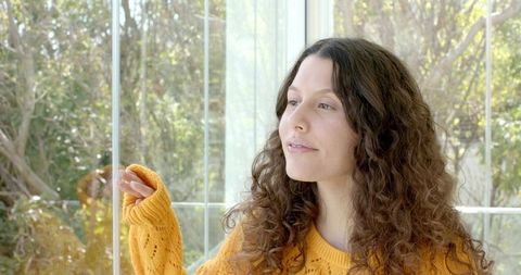 Contemplative woman in mustard knit sweater touching glass wall watching sunlit garden