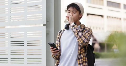 Asian young man drinking coffee and checking smartphone while commuting on urban sidewalk