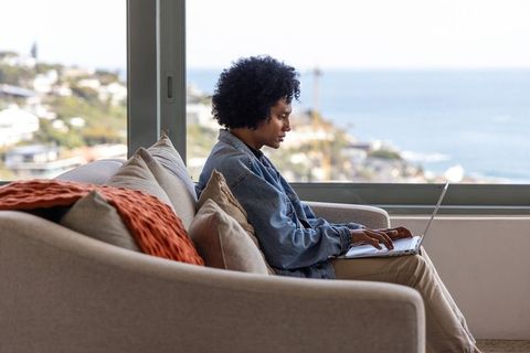 African american man working remotely on laptop on coastal sofa with ocean view