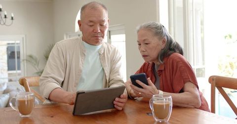 Mature couple sharing digital devices at sunlit kitchen table with coffee, learning