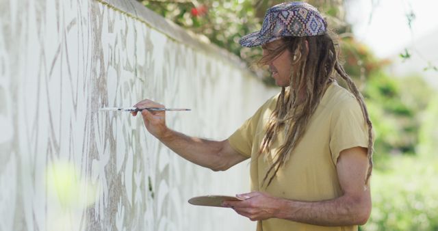 Man with long hair and cap painting white mural on large wall outdoors. Use for artwork themes, urban creativity, contemporary art projects, public spaces revitalization, artistic expressions.
