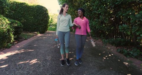 Two Friends Walking Together in Sunlit Park with Water Bottles
