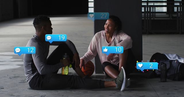 Young athletic couple sitting on floor, having casual conversation after workout session, receiving social media notifications, fitness goal discussion, perfect for use in social media marketing, fitness and wellness campaigns, relationship blogs, or healthy lifestyle promotions.