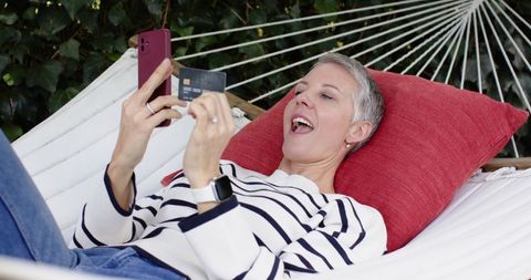 Senior woman relaxing in hammock using smartphone and credit card for online shopping