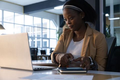 African american woman working at laptop in modern office pointing with pen wearing tan blazer