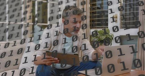 Middle-aged man working remotely on laptop and tablet with binary code overlay