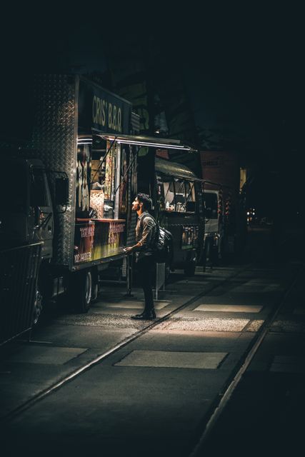 Young man is standing in front of a food truck at night. Street scene features rows of food trucks, with a focus on the illuminated menu. Perfect for concepts related to urban lifestyle, nightlife activities, culinary adventures, and street food culture.