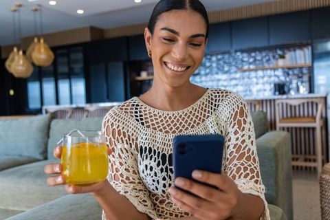 African woman smiling while scrolling smartphone and holding orange juice in modern lounge
