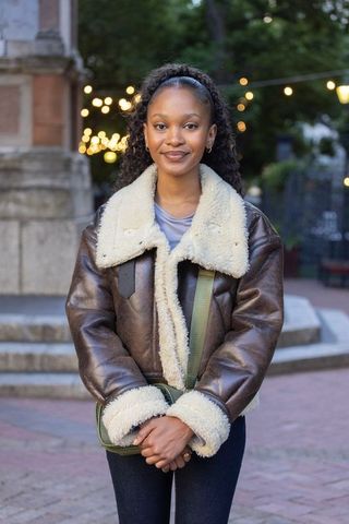 African woman wearing shearling aviator jacket in urban twilight plaza with crossbody bag