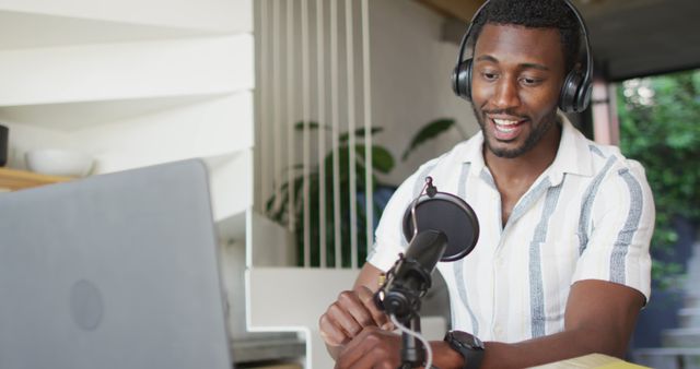 Young African American man recording a podcast at home, sitting at desk with microphone and laptop, headphones on. Ideal for use in blogs and articles on podcasting, digital content creation, remote work, and technology in home office settings.