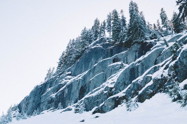 Snow-covered mountain cliff adorned with pine trees perfect for nature and wilderness themes. Useful for winter sports promotions, travel blogs, outdoor adventure advertising, holiday postcards, and winter scenery collections.
