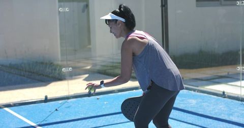 Bending woman reaching for pickleball on sunlit outdoor blue court with paddle and visor