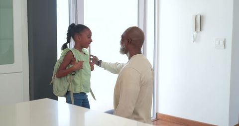 Kneeling father fastening backpack strap while smiling daughter at bright entryway