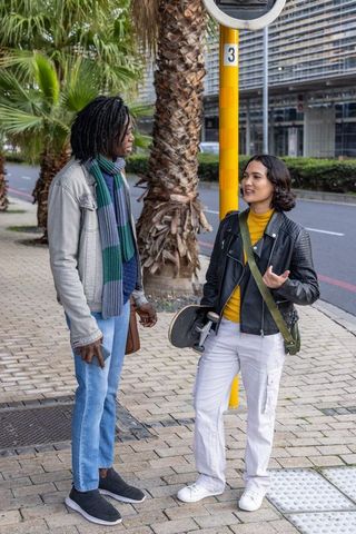 Young duo chatting at bus stop holding skateboard on palm-lined urban sidewalk