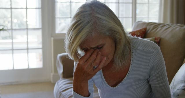 Senior woman showing signs of stress and anxiety while sitting in her living room. Useful for depicting emotional strain, mental health issues, or the daily struggles of elderly individuals. Suitable for articles, blogs, and other educational material related to mental health, senior care, and emotional well-being.