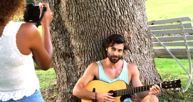 Man sitting against tree trunk, playing acoustic guitar while woman photographs moment with a camera. Perfect for themes of outdoor leisure, music, creativity, young couple activities, and summer relaxation.