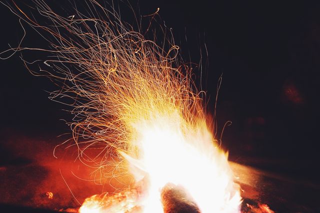 Close-up of a mesmerizing campfire at night with sparks flying in various directions, creating a captivating display. Ideal for use in outdoor adventure promotions, camping equipment advertisements, or as a representation of warmth and togetherness during evening gatherings in nature.
