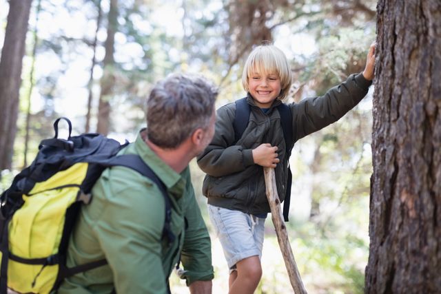 Cheerful boy looking at father while standing by tree trunk in forest