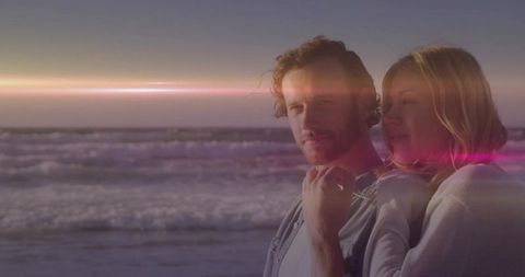 Romantic couple watching sunset on beach with pink lens flare, breaking waves, warm golden glow
