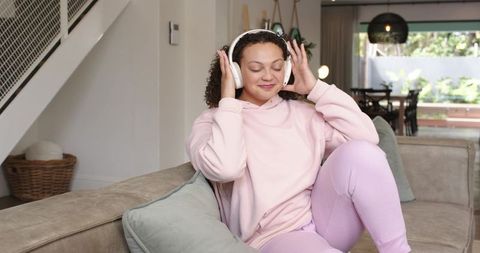 African american woman listening to music on wireless headphones, relaxing on pastel sofa