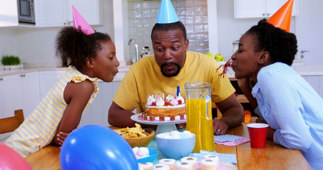 Family celebrating birthday in kitchen with father and daughters wearing party hats blowing out candle on cake. Great for advertisements, blogs, social media posts about family events, birthdays, celebrations, and joyful moments.