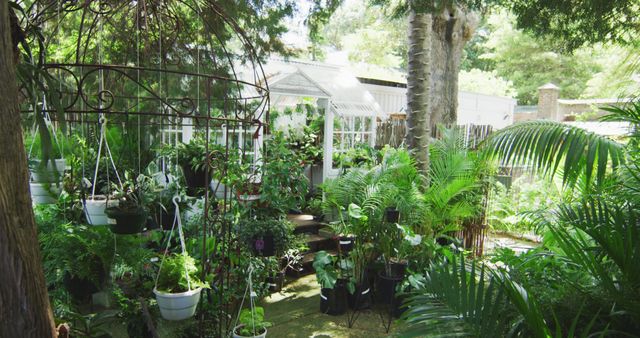 Front view of a gazebo with hanging plant pots on it, surrounded by green plants and trees, with a greenhouse in the background, in slow motion