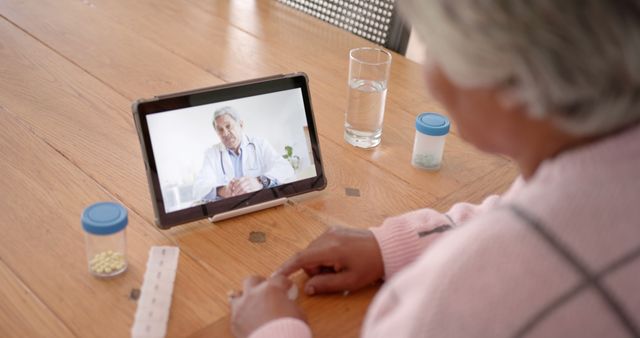 Senior woman having a video chat with a doctor for a medical consultation on a tablet. Pills and water on table. Perfect for articles on telehealth, online medical services, elderly healthcare management, digital health advancement.