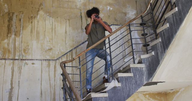 Male photographer using retro camera in aged, abandoned building, descending concrete staircase stained with graffiti. Suitable for themes of urban exploration, adventure, hobbies, and curiosity, or use in illustrating concepts of decay and history.