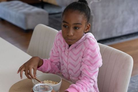 School-age african girl eating cereal in pink fleece at home, cozy natural light morning