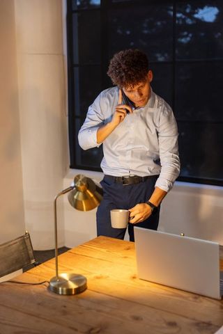 Young professional talking on phone while holding coffee at evening desk with laptop