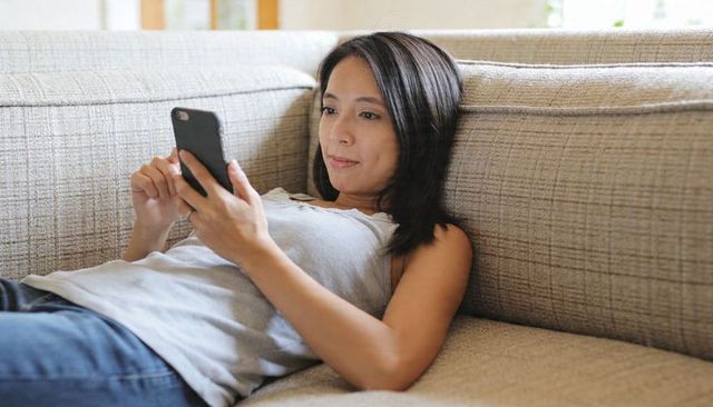 Relaxing woman lounging on beige sofa using smartphone for browsing, texting, shopping