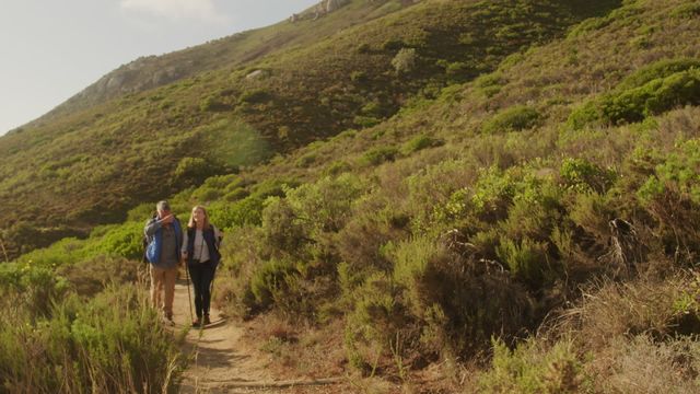 This serene image features a senior Caucasian couple trekking through a scenic mountainous area with backpacks and Nordic walking sticks. Ideal for use in adventure and lifestyle content promoting outdoor activities among the elderly. It showcases themes of exploration, fitness, and companionship in nature.