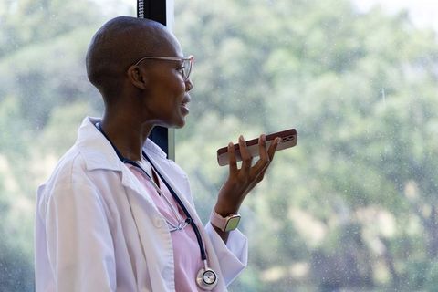 African female doctor speaking into smartphone for telemedicine consultation by window