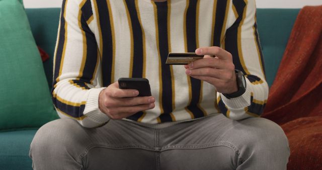 Man wearing casual striped sweater sitting on couch holding credit card and smartphone, suggesting online shopping or mobile banking. Perfect for illustrating concepts related to e-commerce, digital payments, online security, and modern lifestyle.