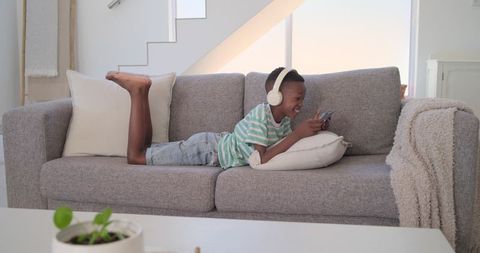 Preteen lounging on gray sofa wearing white headphones using smartphone in bright living room