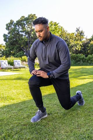 Man performing forward lunge on sunlit backyard lawn wearing quarter-zip, joggers, smartwatch